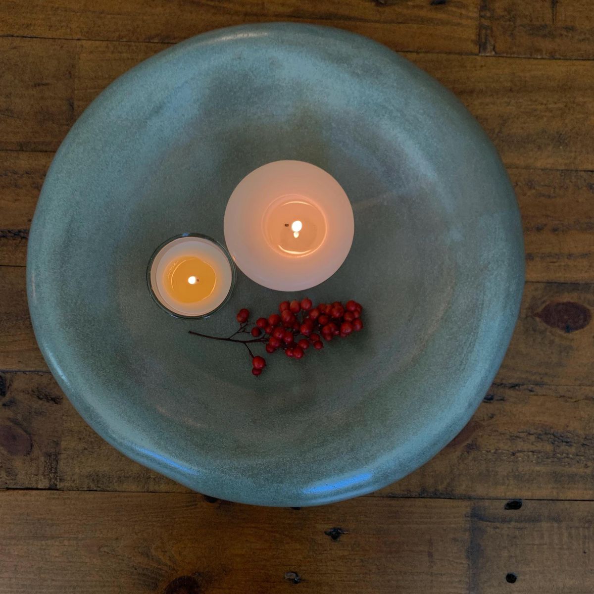 Two lit candles and red berries in a teal ceramic bowl on a wooden surface