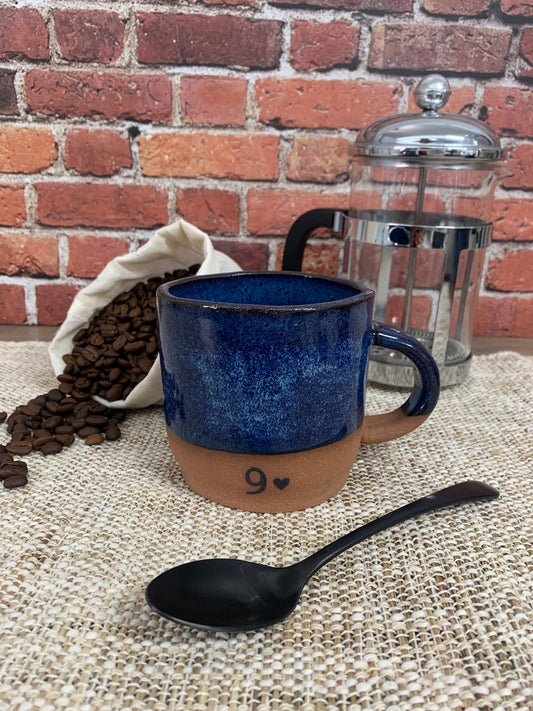 a blue handmade pottery mug for anniversary gift with a nine and heart printed on front, next to a spoon, a bag of coffee beans and french press with a rick background