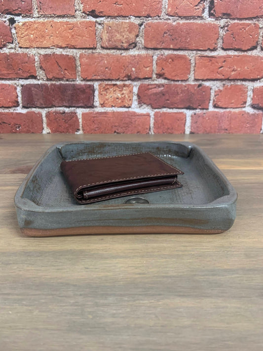Brown leather wallet on a gray pottery catchall tray stamped 9 and heart with a brick wall background