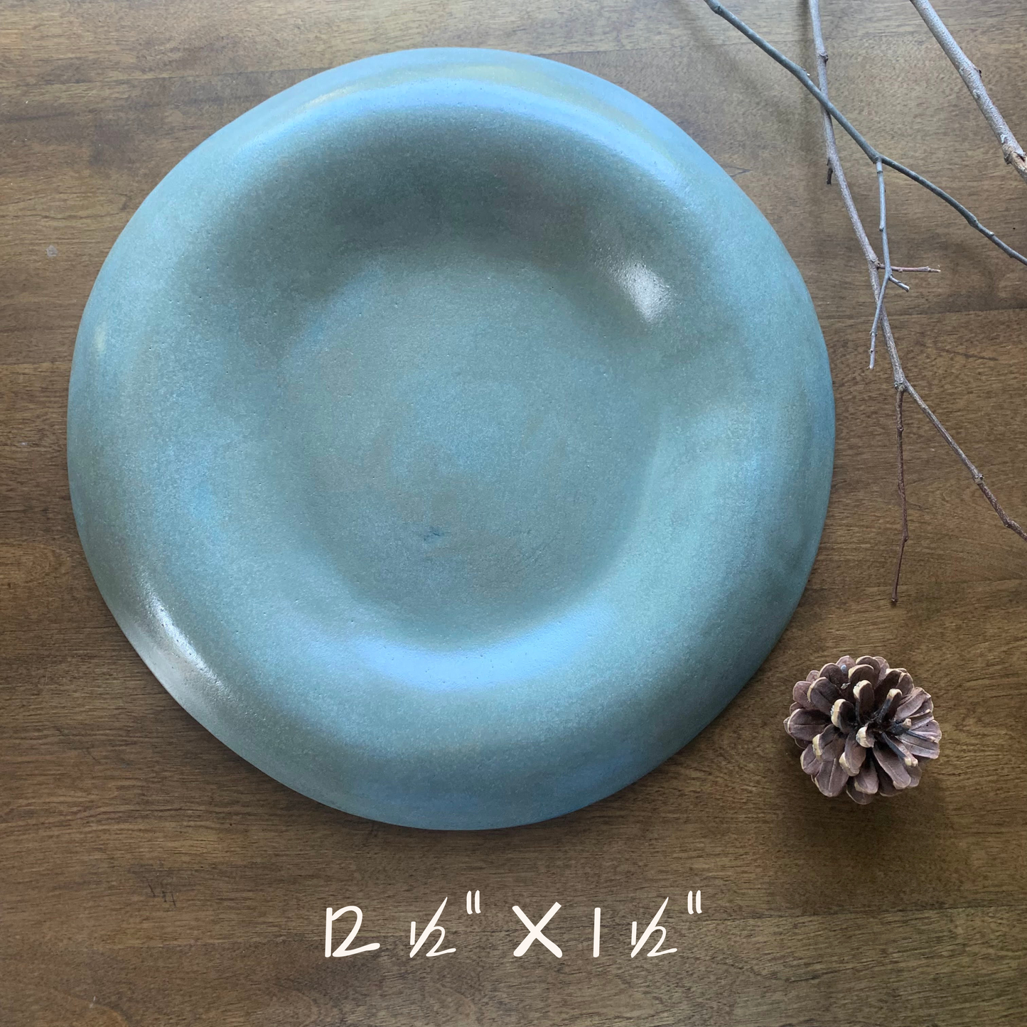 Blue-green ceramic plate on a wooden surface with a pine cone text with dimensions 12.5" x 1.5"