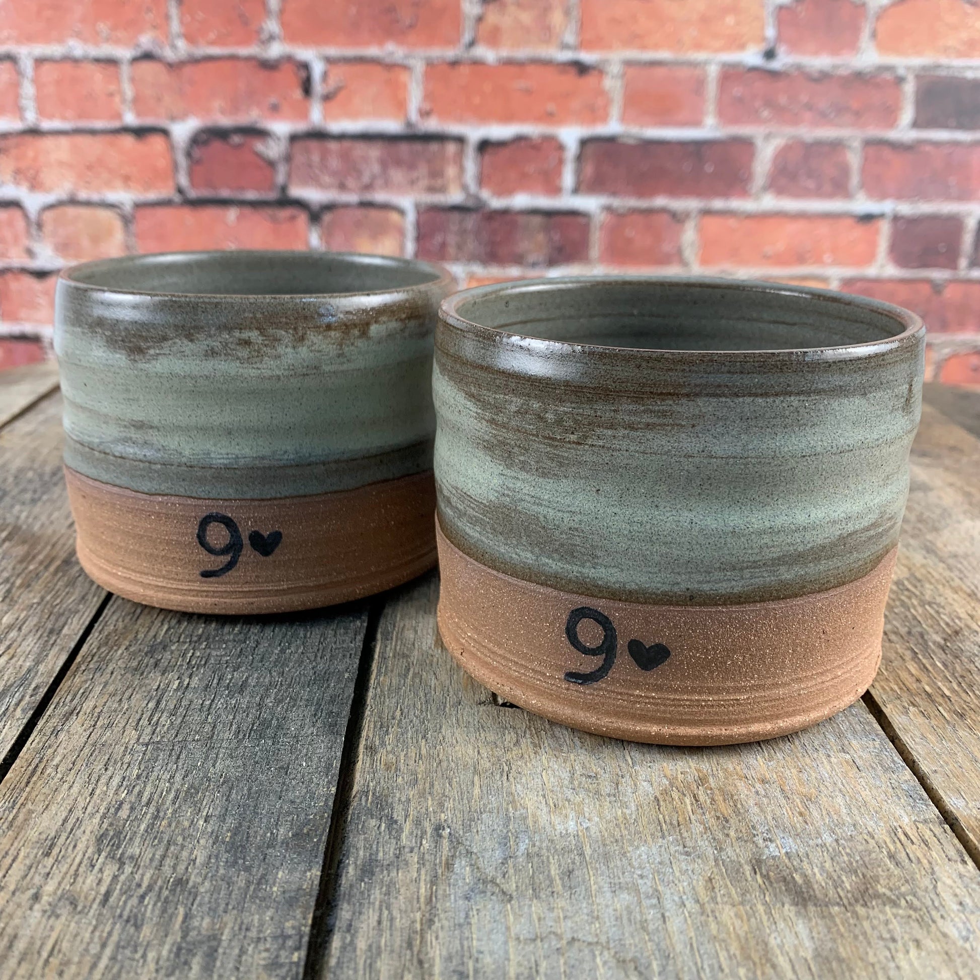 Two pottery tumblers with brown and gray glaze on a wooden surface with a brick wall background.