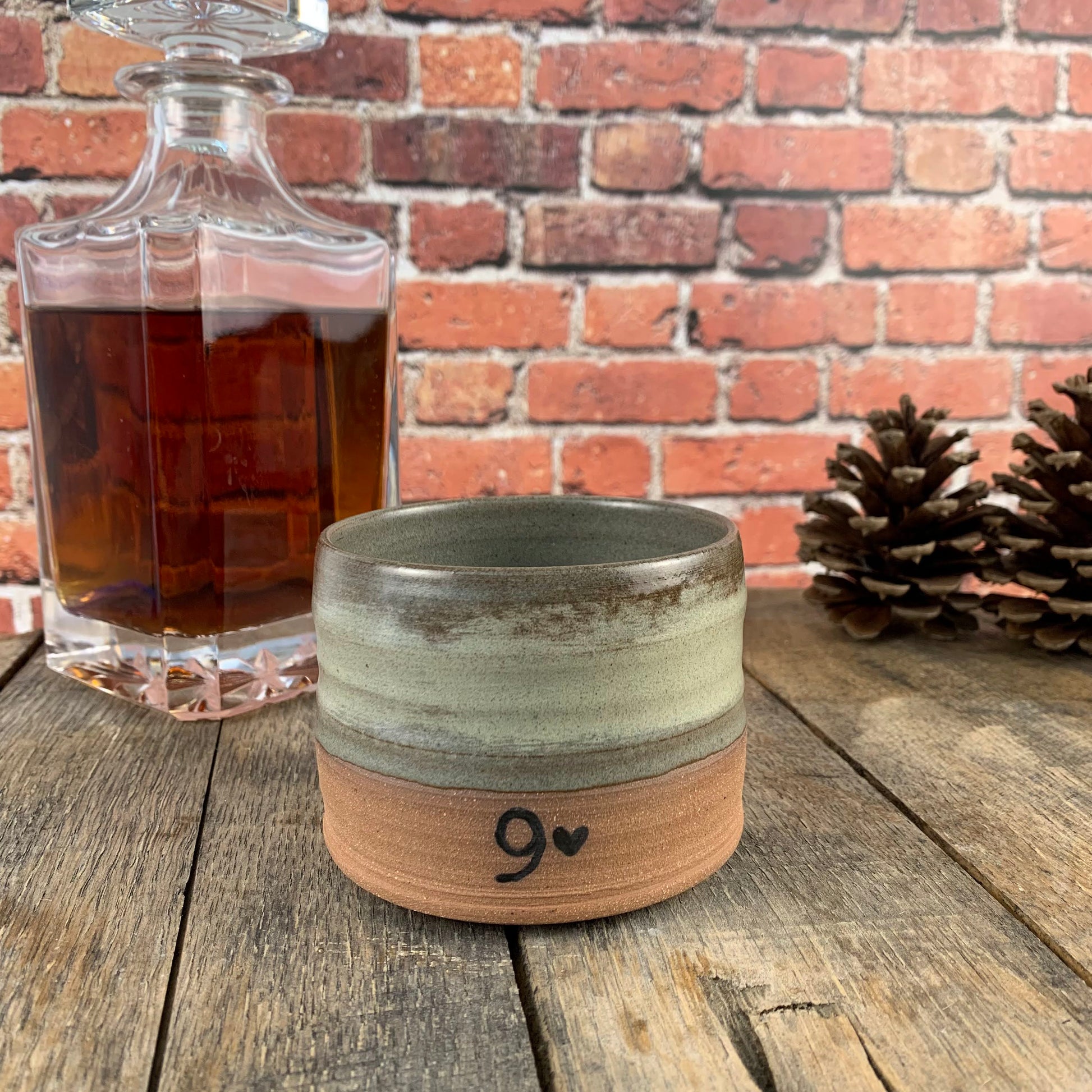 Gray ceramic pottery cup on wood surface next to bottle of whiskey and pine cones