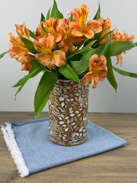 Bouquet of orange flowers in a decorative handmade vase with leaf designs on a wooden surface with a blue cloth.