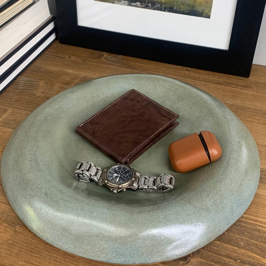 Brown leather wallet and watch on a green catchall entryway dish with a framed picture in the background