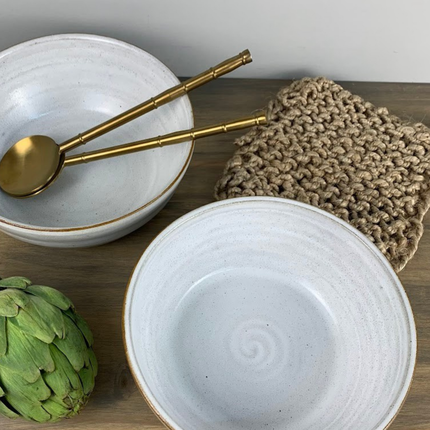 Two white ceramic bowls with gold serving spoons inside on a wooden surface, accompanied by a green artichoke and a beige knitted cloth.