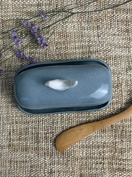 Blue ceramic butter dish with a white interior on a textured fabric background with a wooden spoon and lavender.