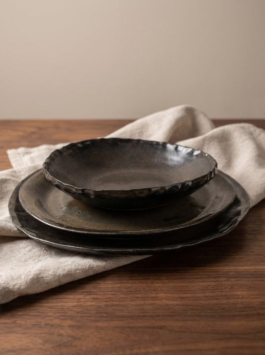 Set of brown hand formed ceramic plates and bowl on a wooden table with a beige napkin.