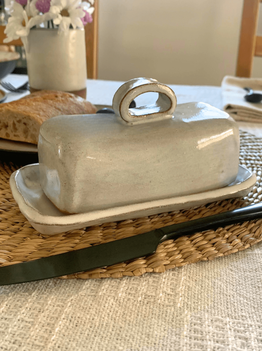 Butter dish with lid on a woven tray on a table with bread and flowers in the background