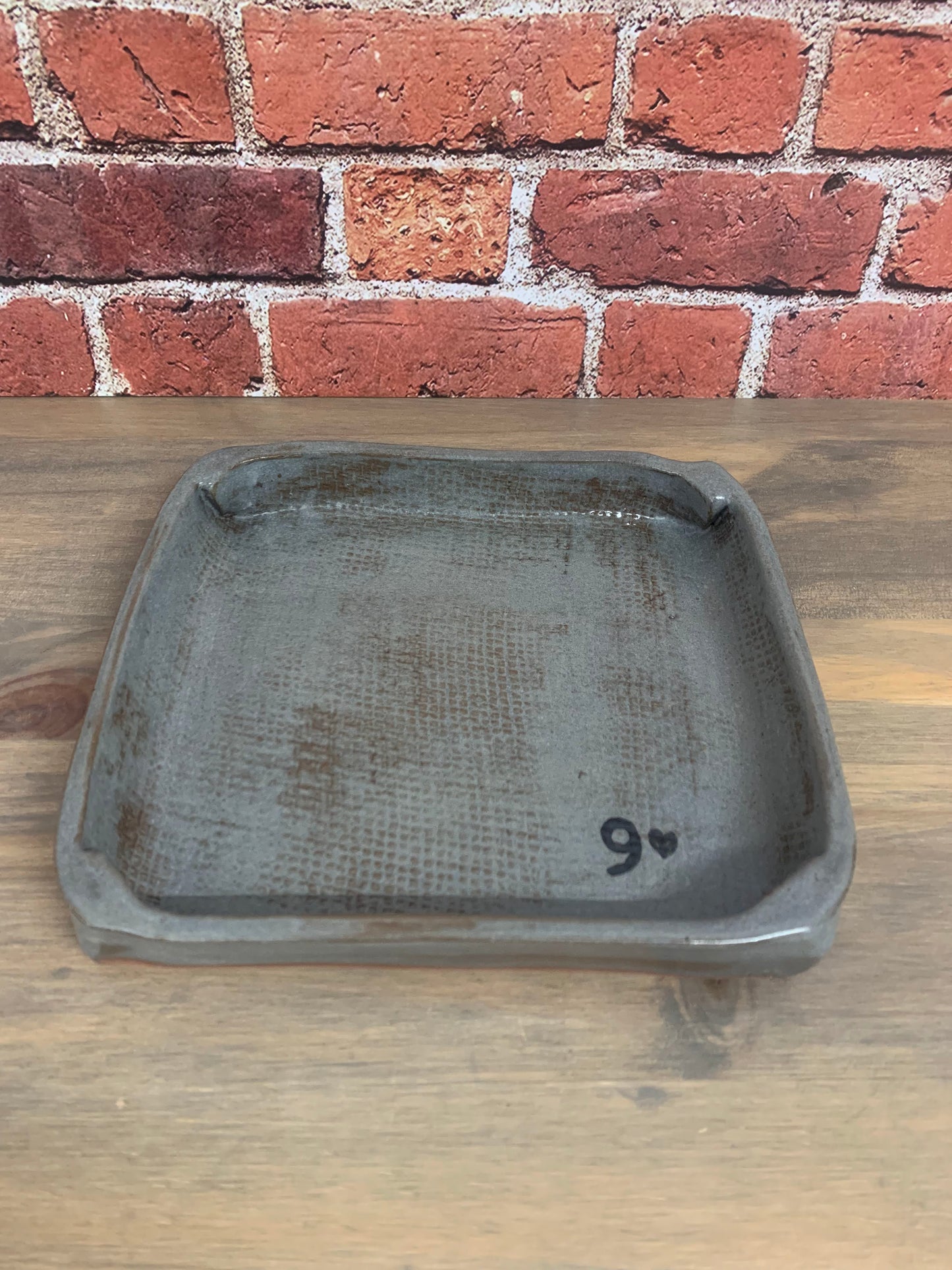 handmade rustic pottery tray with '9' on a wooden surface next to a brick wall