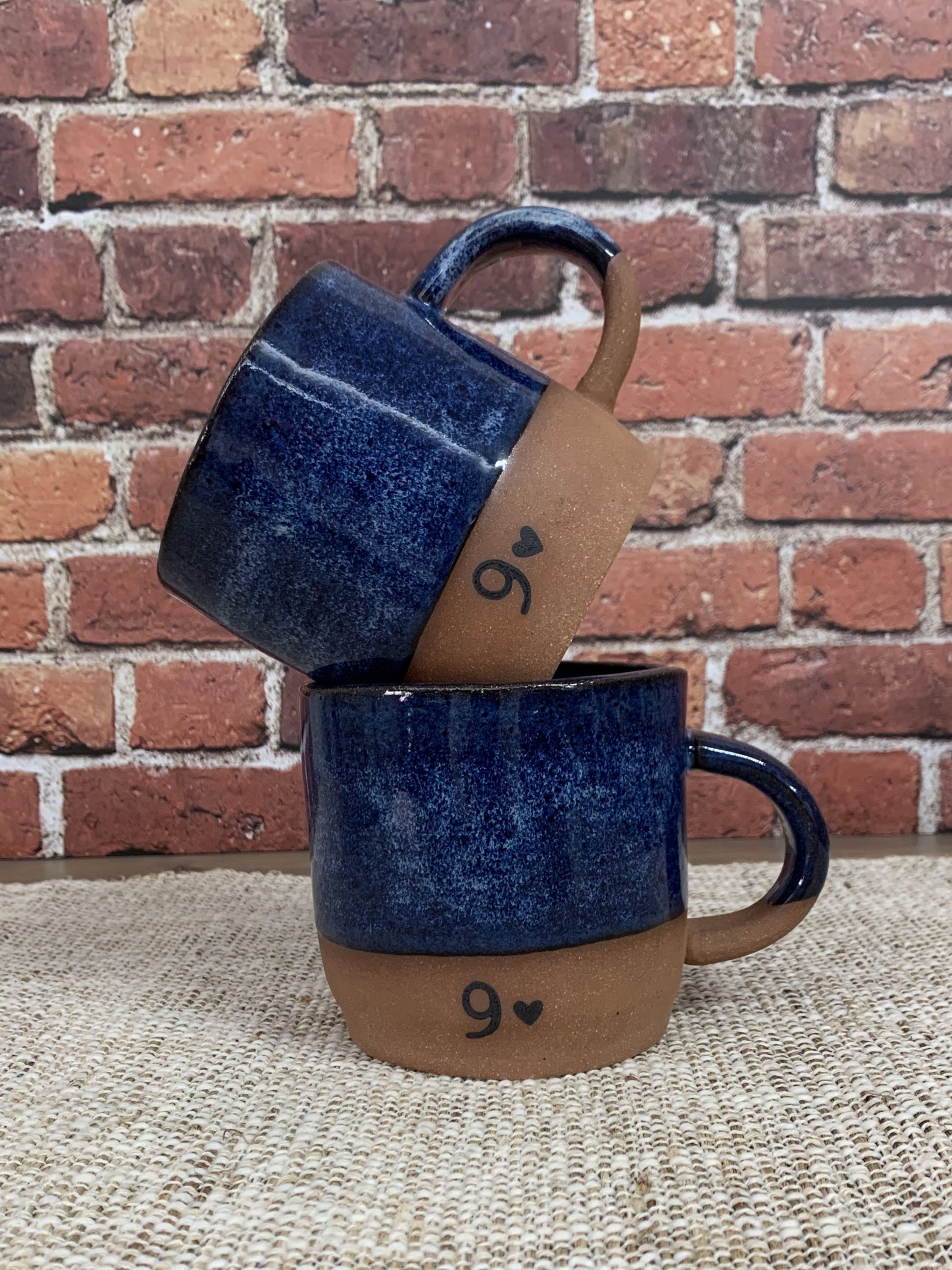Two blue ceramic 9th anniversary  mugs with brown bases stamped with a nine and a heart and handles against a brick wall.