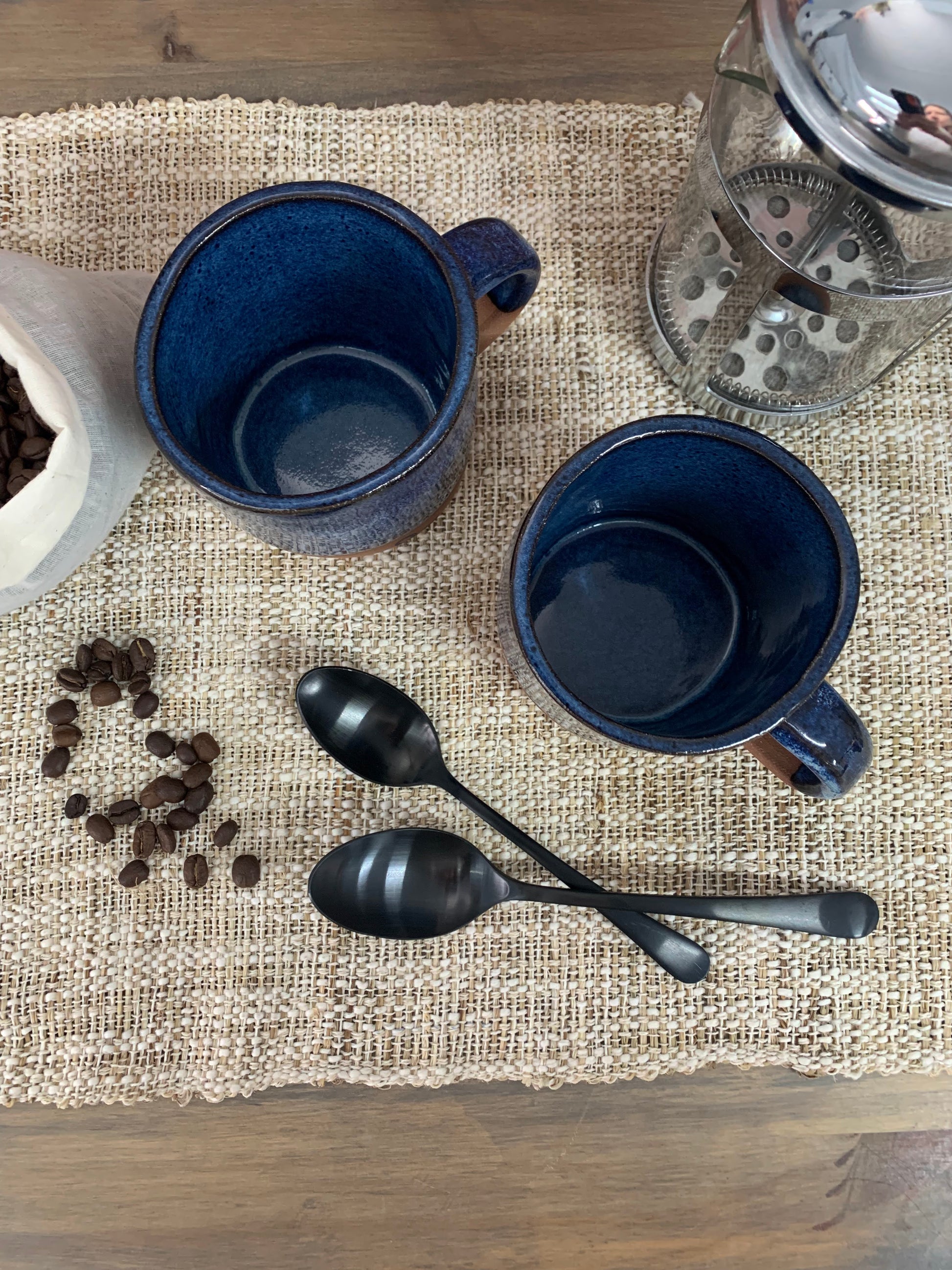 Two blue anniversary mugs, two black spoons, and coffee beans on a textured surface.