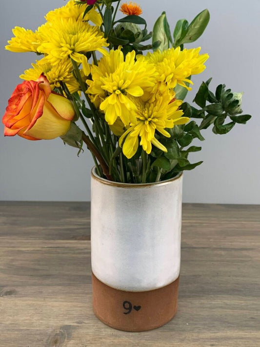 Bouquet of yellow flowers and a red rose in a white handmade pottery vase with a brown base on a wooden surface.