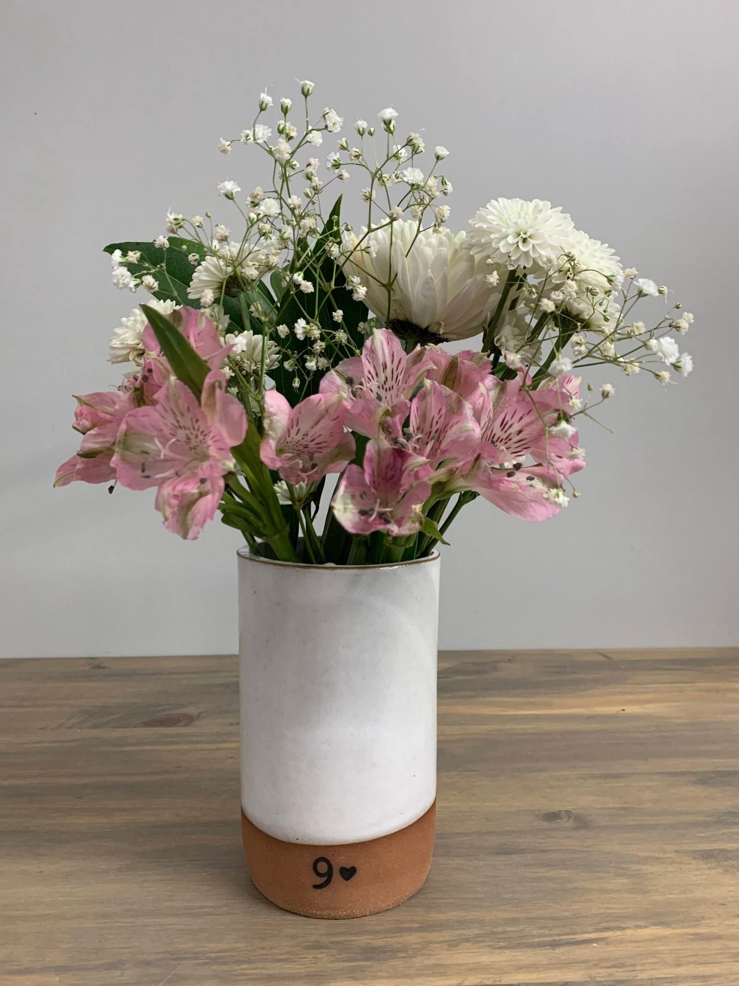 Bouquet of pink and white flowers in a white vase with a brown base on a wooden surface.