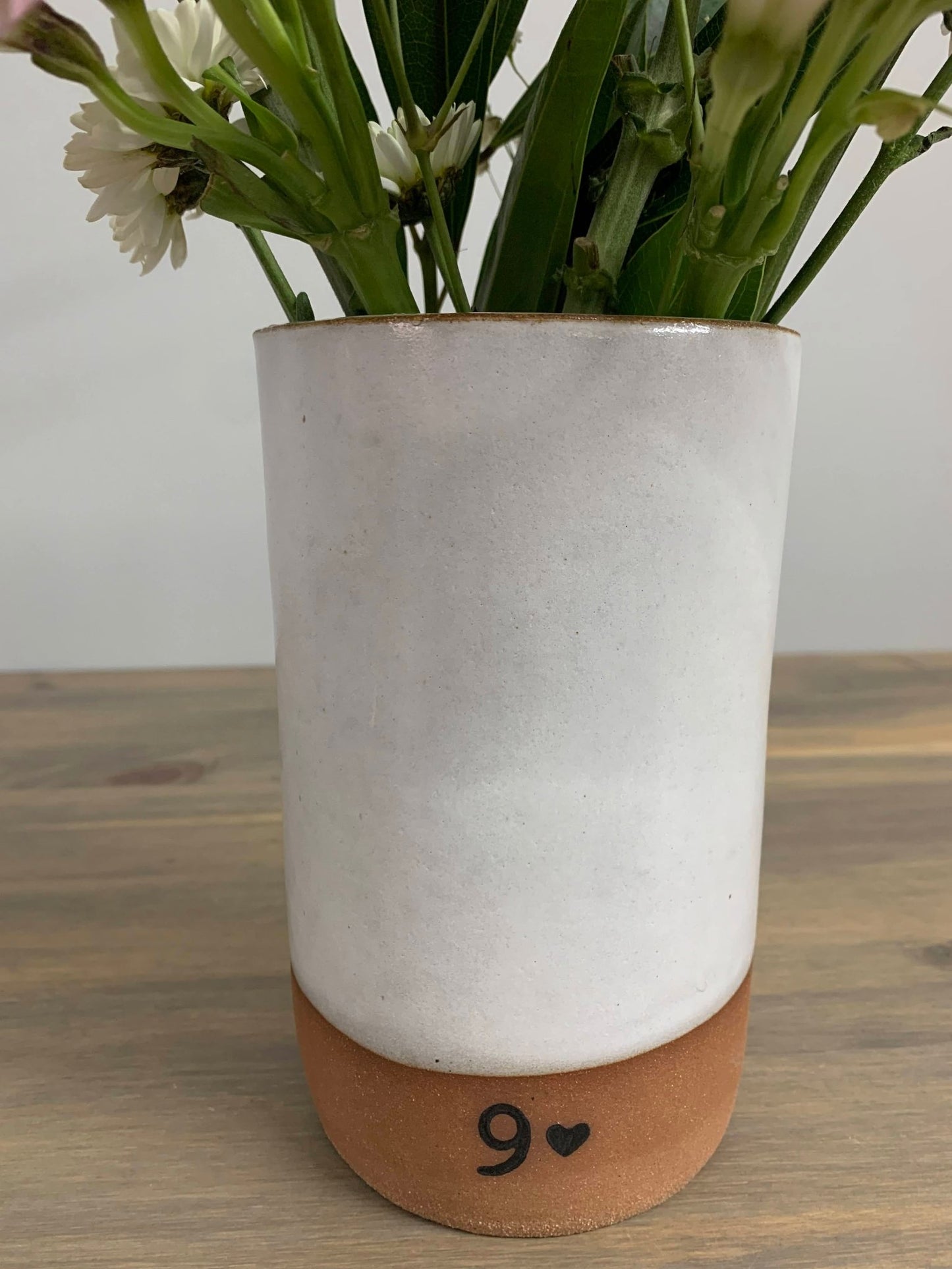 White handmade pottery vase with a brown base with a 9 on it on a wooden surface, containing greenery and white flowers.