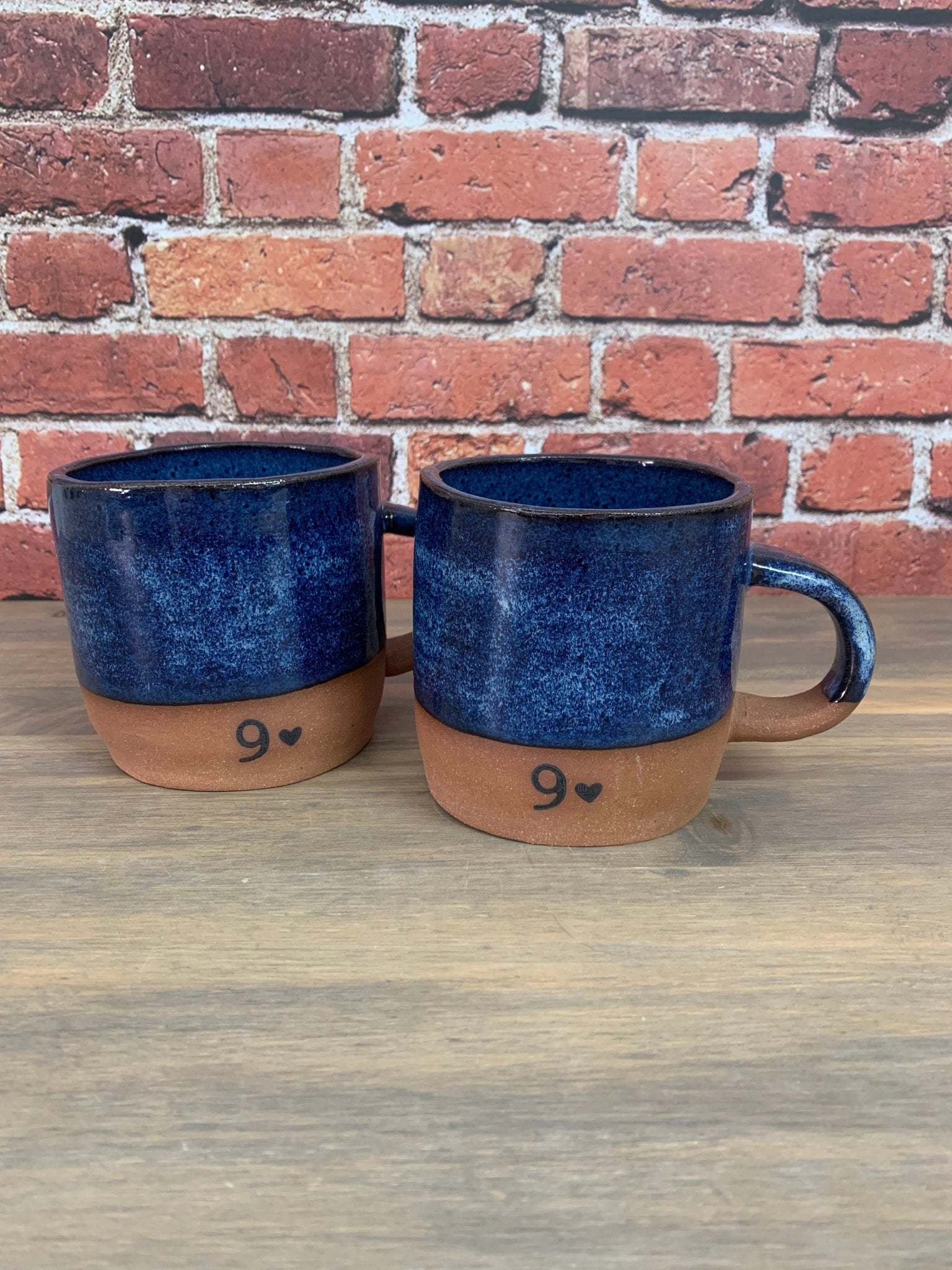 Two blue pottery anniversary mugs with brown bases and number 9 printed on front on a wooden surface against a brick wall.