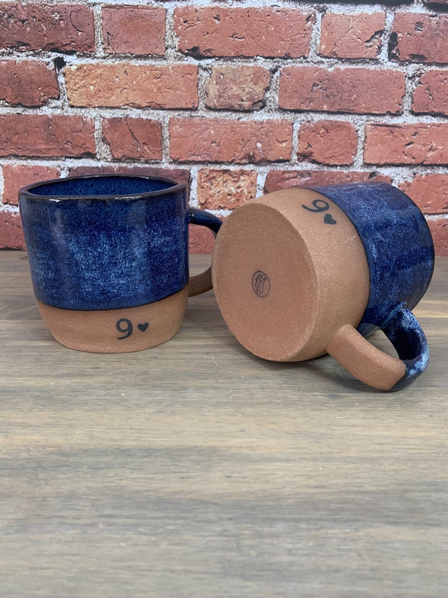Two ceramic pottery anniversary mugs with blue and brown colors and number 9 printed on base, on a wooden surface with a brick wall background.