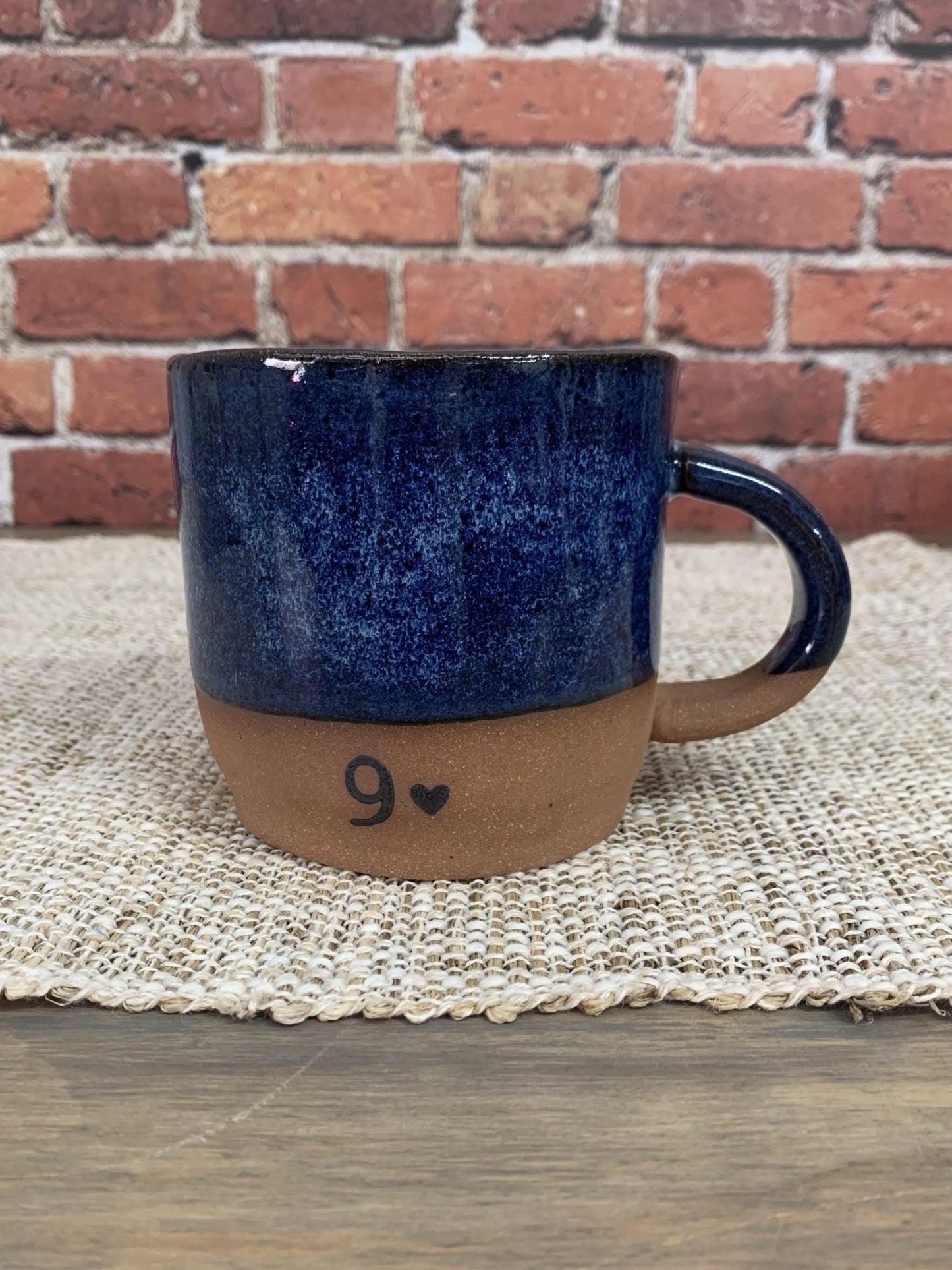 Blue ceramic mug with brown base and number 9 on a woven mat against a brick wall.
