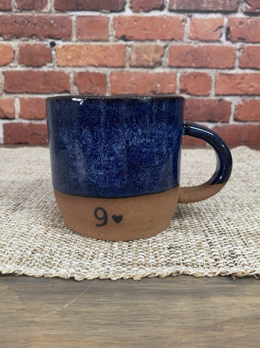 Blue ceramic mug with brown base and number 9 on a woven mat against a brick wall.