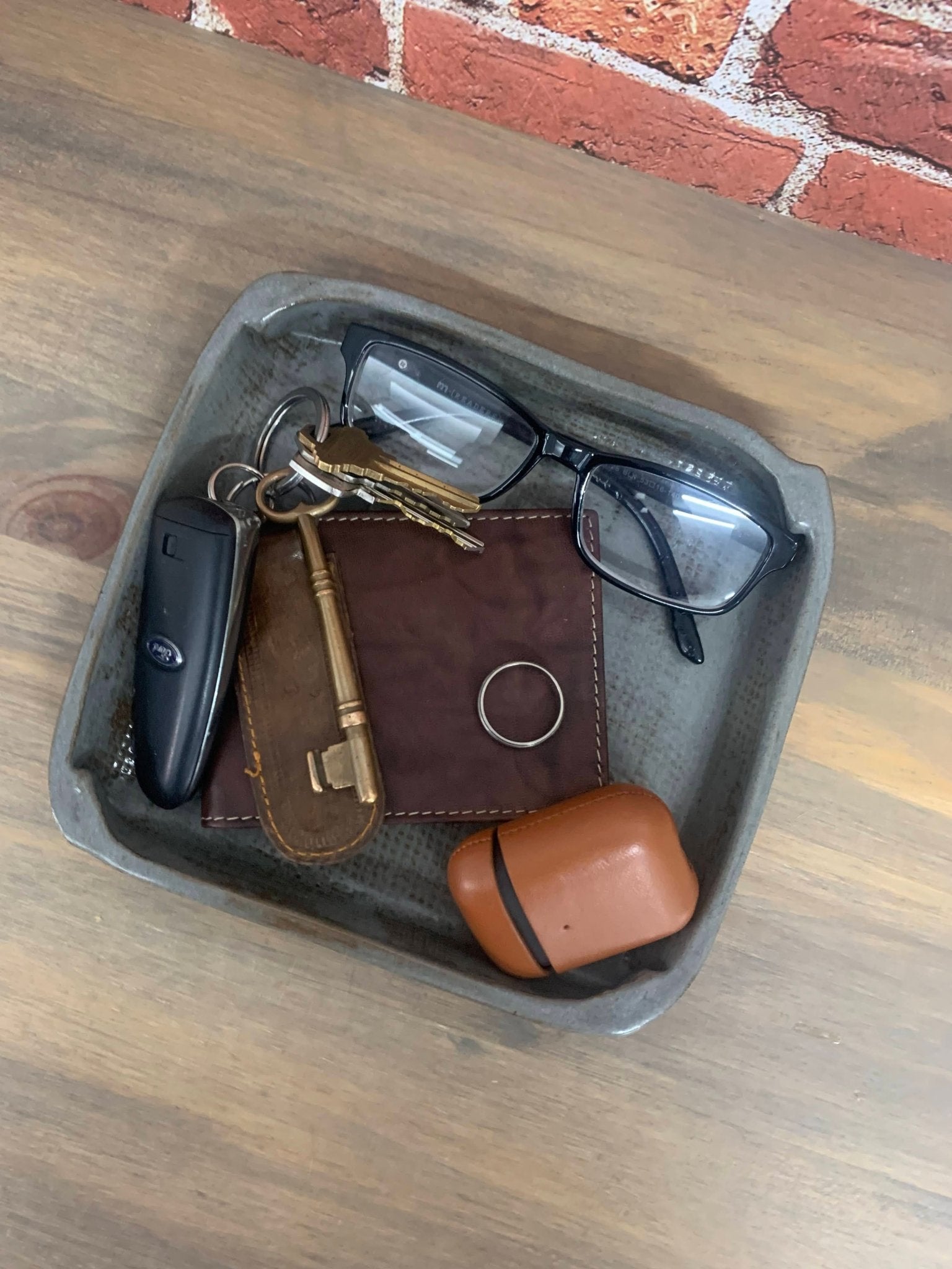pottery Tray with keys, wallet, glasses, and earbuds on a wooden surface with brick wall background