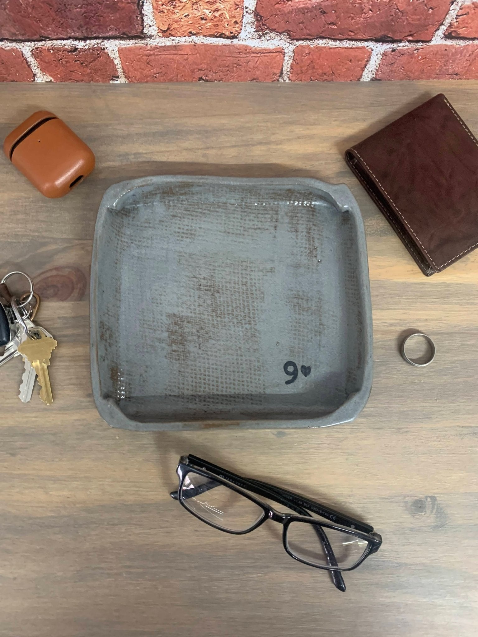 Gray pottery tray with a number 9 and heart holding wallet, glasses, keychain, and ring on wooden surface