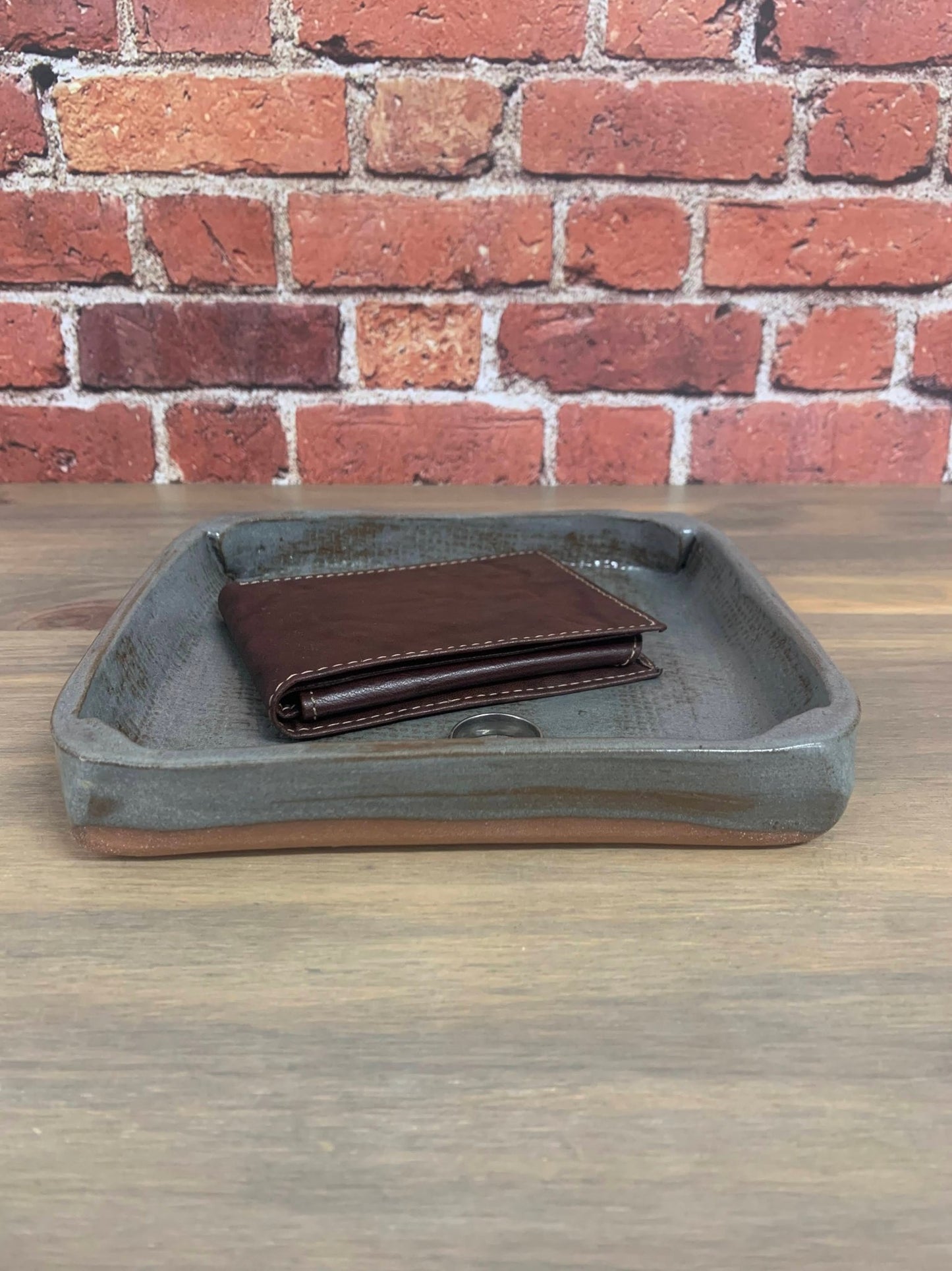 Brown leather wallet on a gray pottery catchall tray stamped 9 and heart with a brick wall background