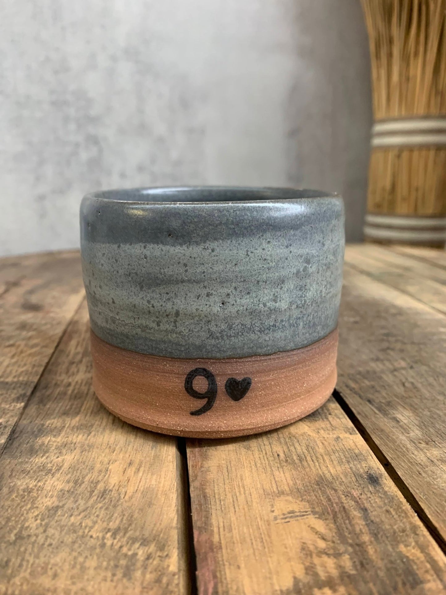 wheel thrown stoneware cup with gray glaze and a number 9 and heart printed close to the bottom, sitting on rustic wood table