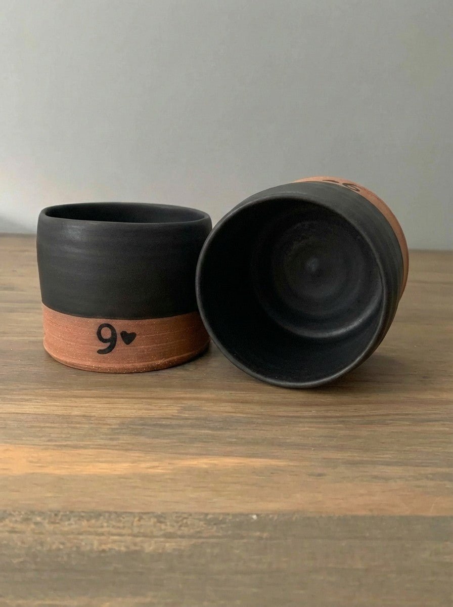 Two black pottery whiskey cups with 9 design on a wooden surface