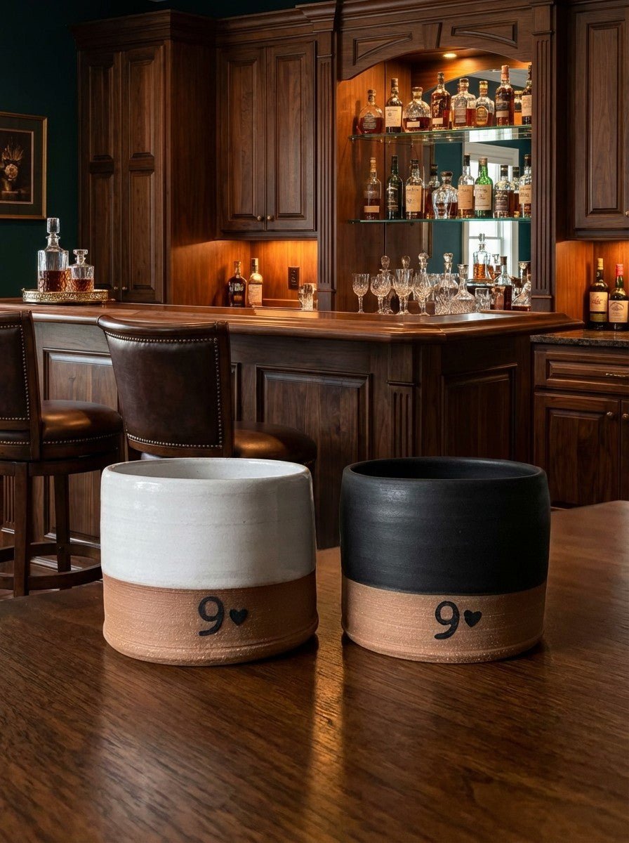 Two pottery whiskey cups with '9' and heart design on a wooden table in a bar setting.