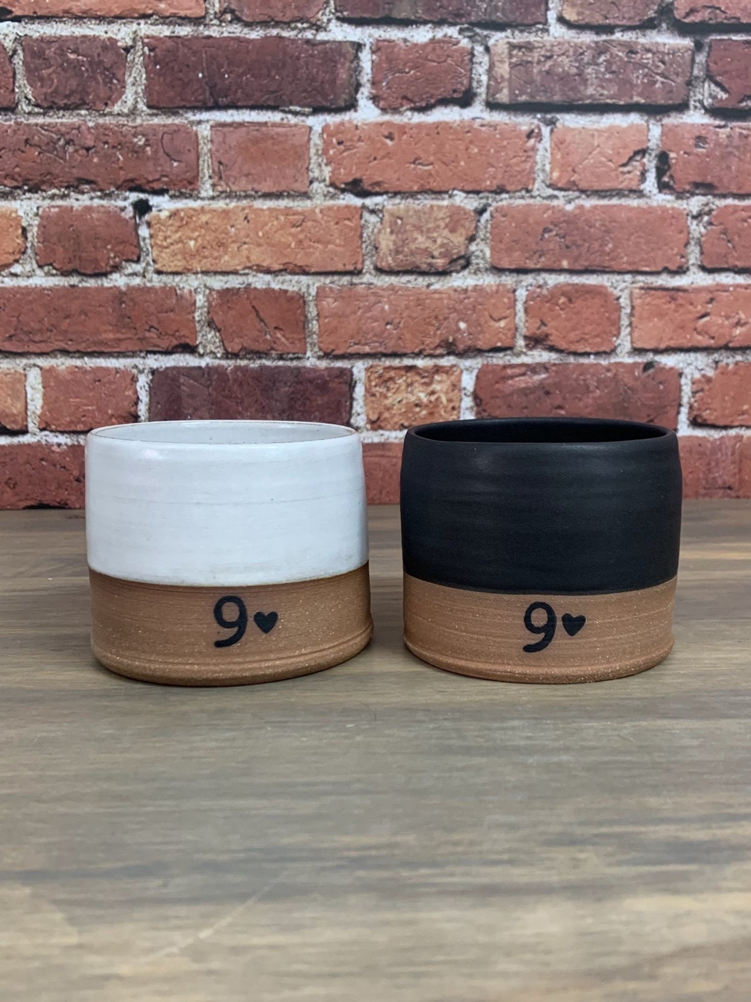 Two pottery tumblers with number 9 and hearts on a wooden surface against a brick wall.