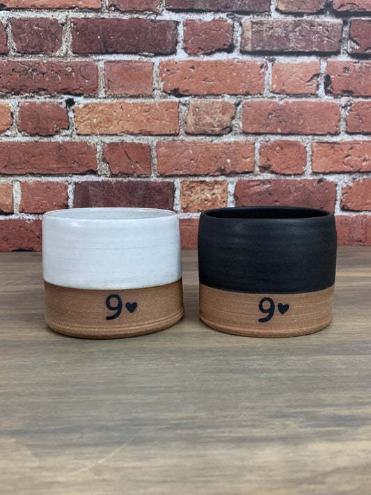 Two pottery tumblers with number 9 and hearts on a wooden surface against a brick wall.