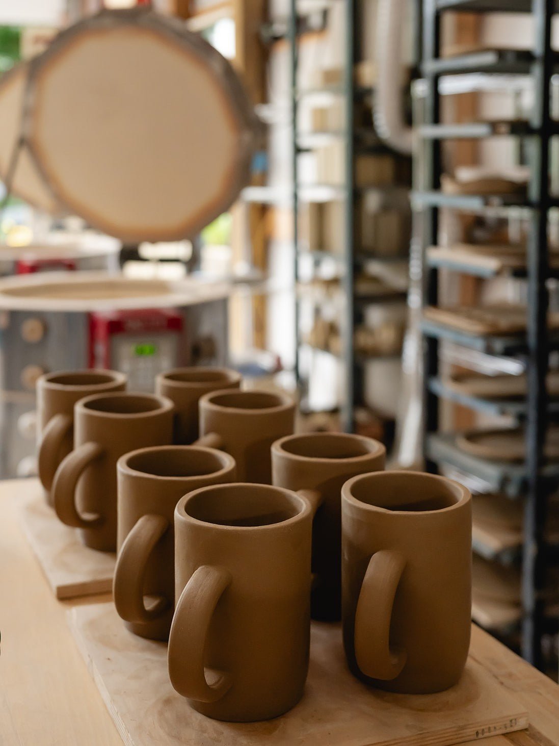 Row of brown ceramic mugs on a wooden surface with a pottery studio in the background.