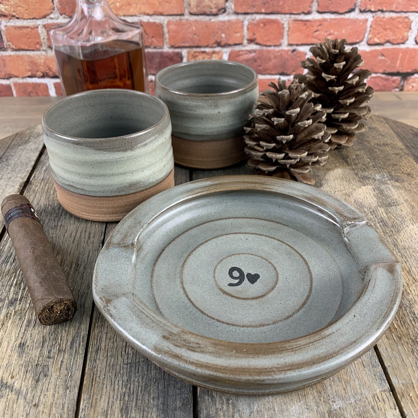 Set of ceramic whiskey tumblers and a cigar ashtray with a 9 and heart design on a wooden surface against a brick wall.
