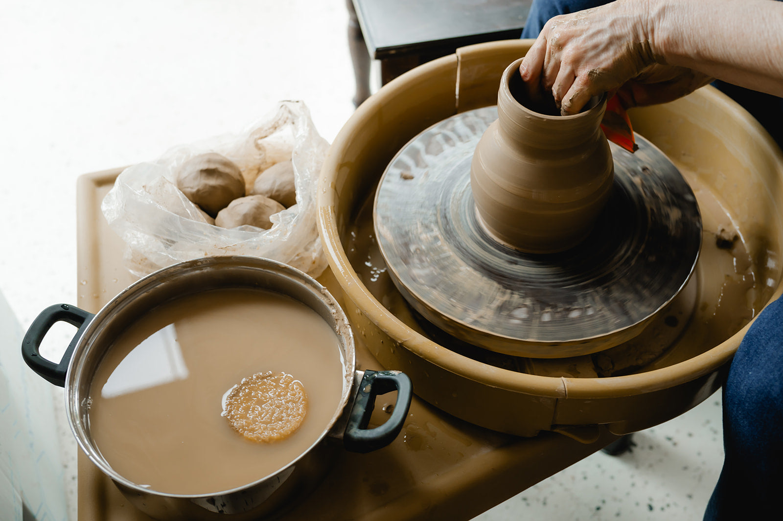 Person working on a potter's wheel with a bowl of water and a bag of clay nearby.