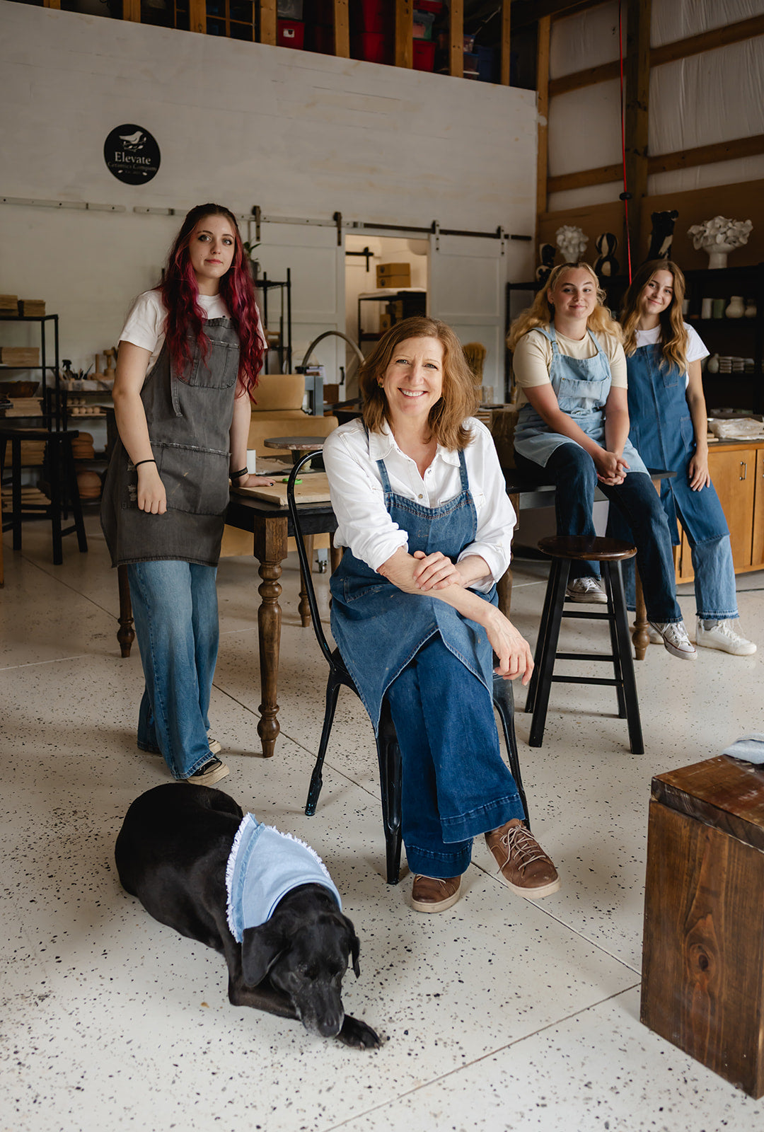 Four women in aprons standing in a workshop with a dog wearing a blue towel.