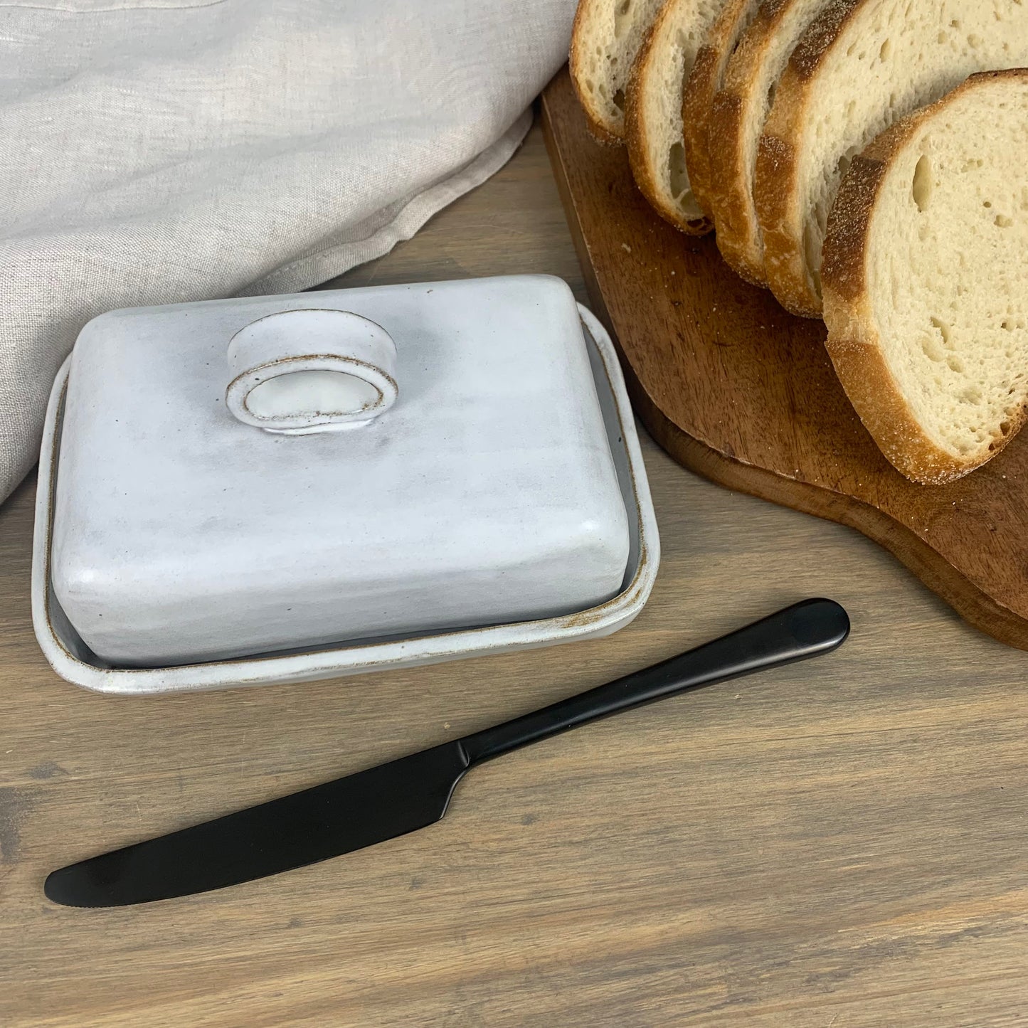 White wide size butter dish with black knife on a wooden surface with sliced bread