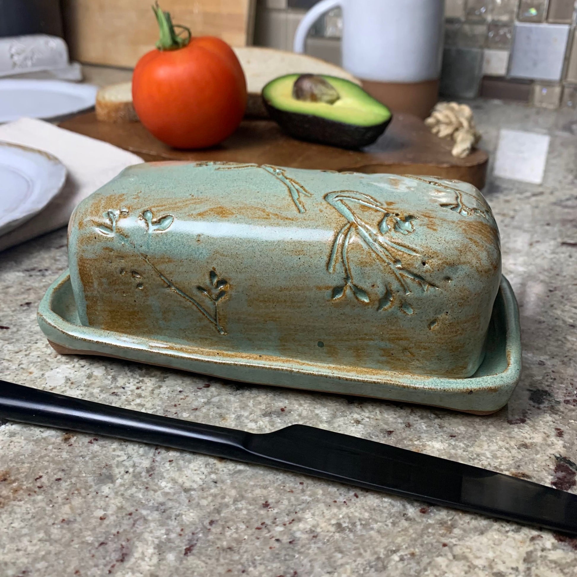 Green artisan ceramic butter dish with decorative elements on a kitchen counter with a knife and fruit in the background.