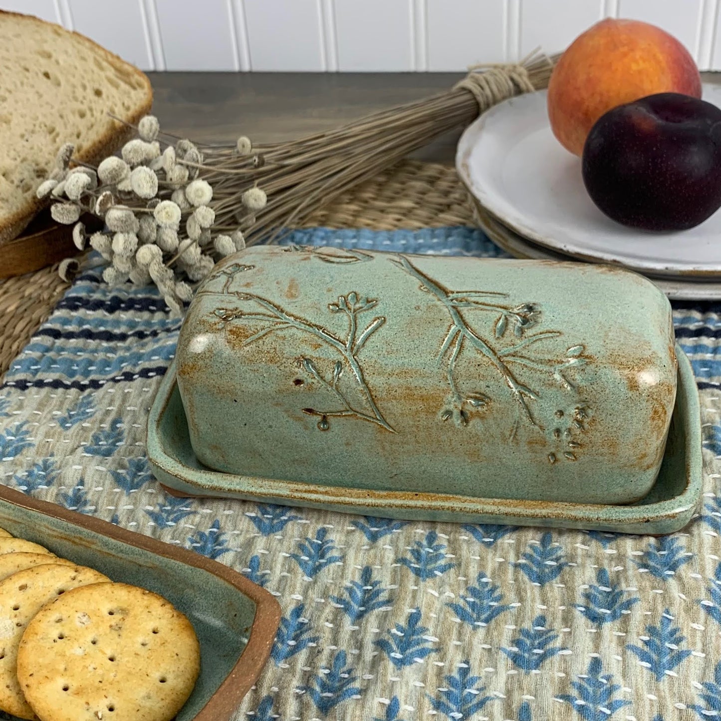 Decorative handmade ceramic butter dish with floral design on a table with bread, crackers, and fruit.