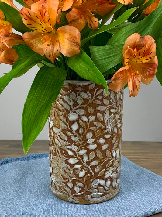 Cylindrical handmade ceramic vase with intricate leaf pattern with bouquet of flowers on a blue cloth