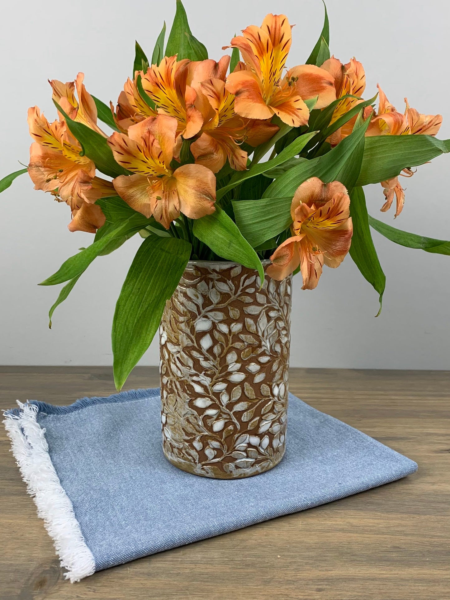 Bouquet of orange flowers in a decorative handmade vase with leaf designs on a wooden surface with a blue cloth.