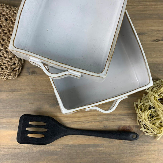Two ceramic baking dishes with white handles on a wooden surface, accompanied by a black spatula and uncooked pasta.