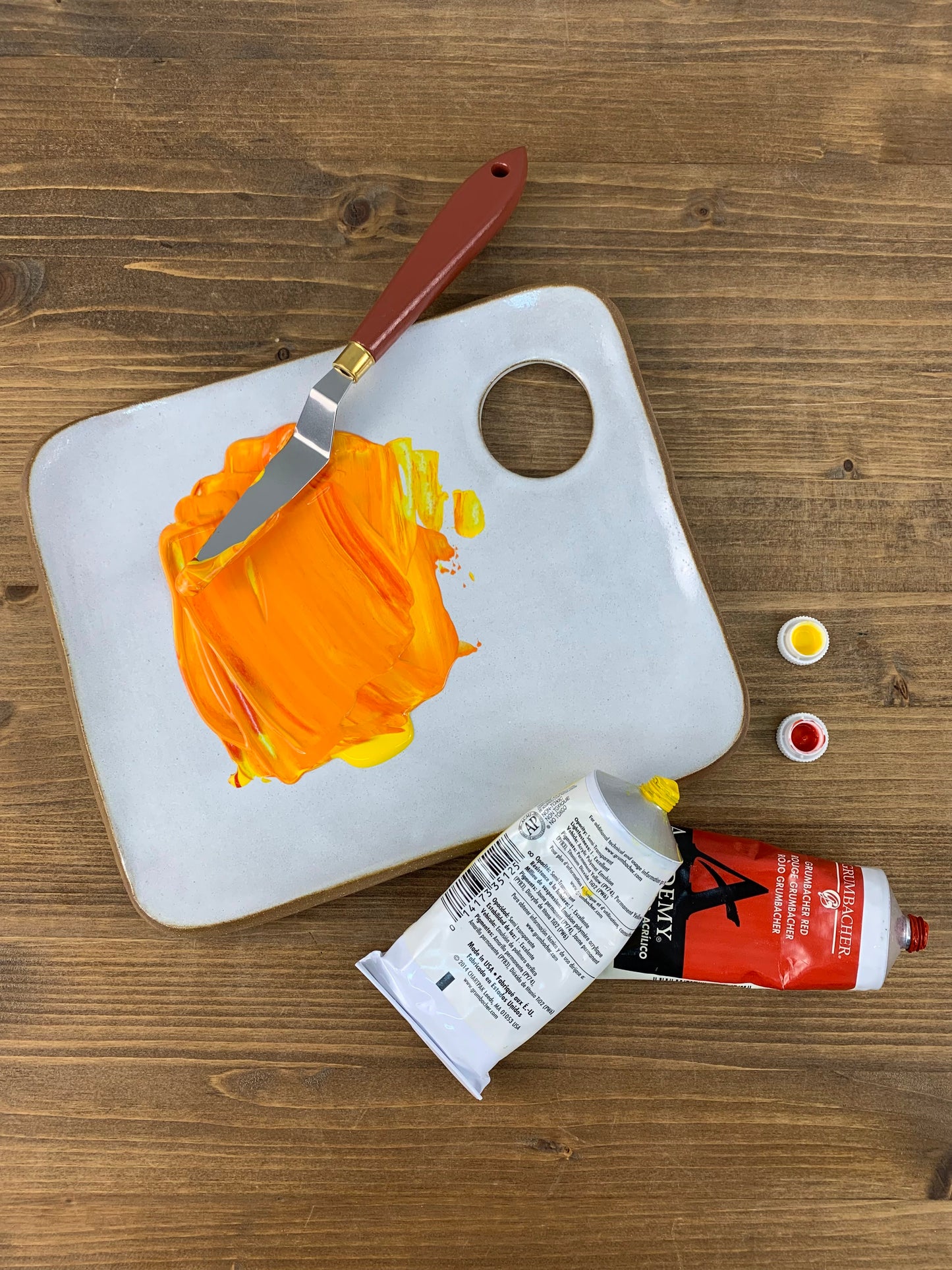 White ceramic paint palette with orange paint, a knife, and art supplies on a wooden surface