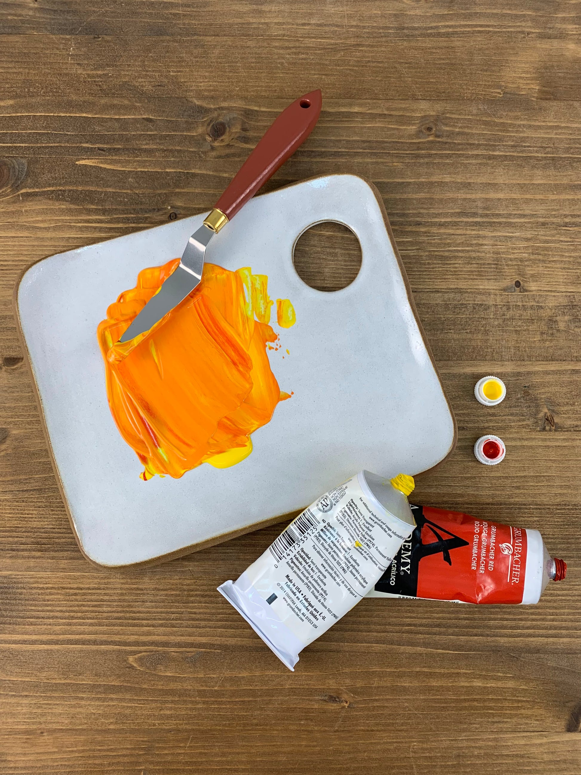 White ceramic paint palette with orange paint, a knife, and art supplies on a wooden surface