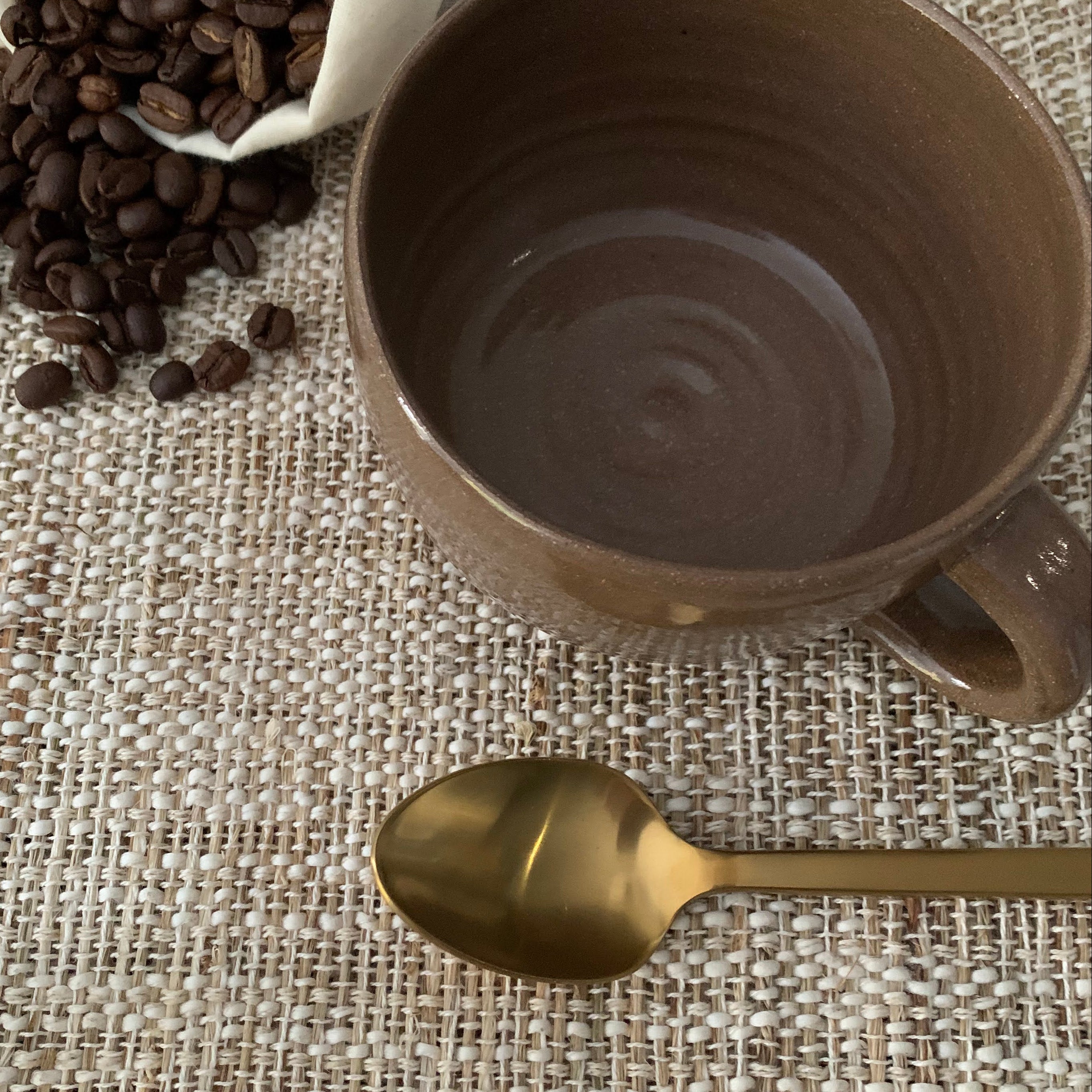Brown ceramic mug, gold spoon, and coffee beans on a textured surface