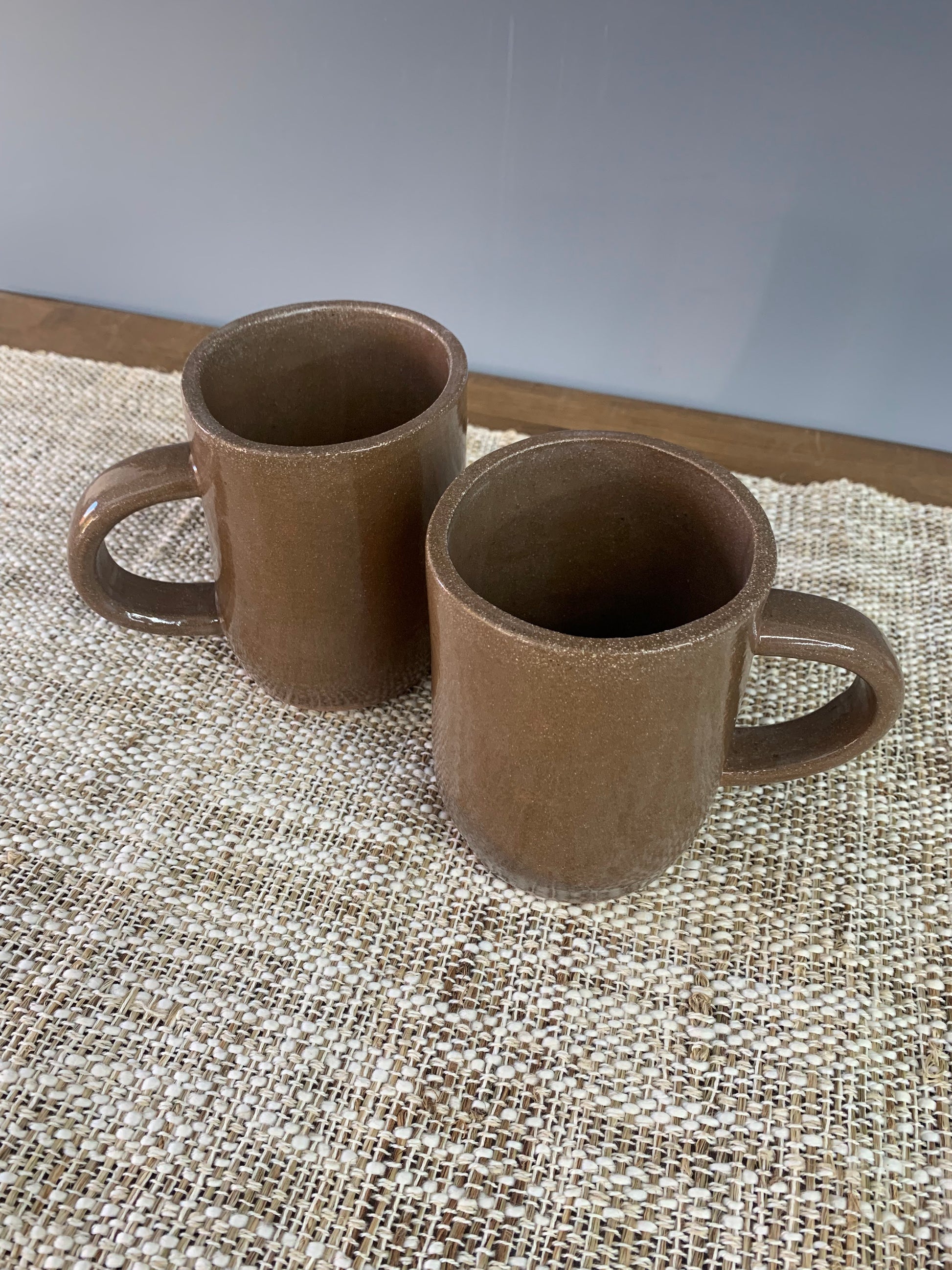 Two brown handcrafted ceramic pottery mugs with handles on a textured surface