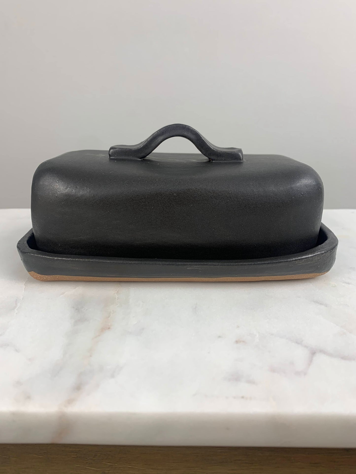 front view of a handcrafted Black ceramic butter dish with lid and simple strap handle on a marble surface