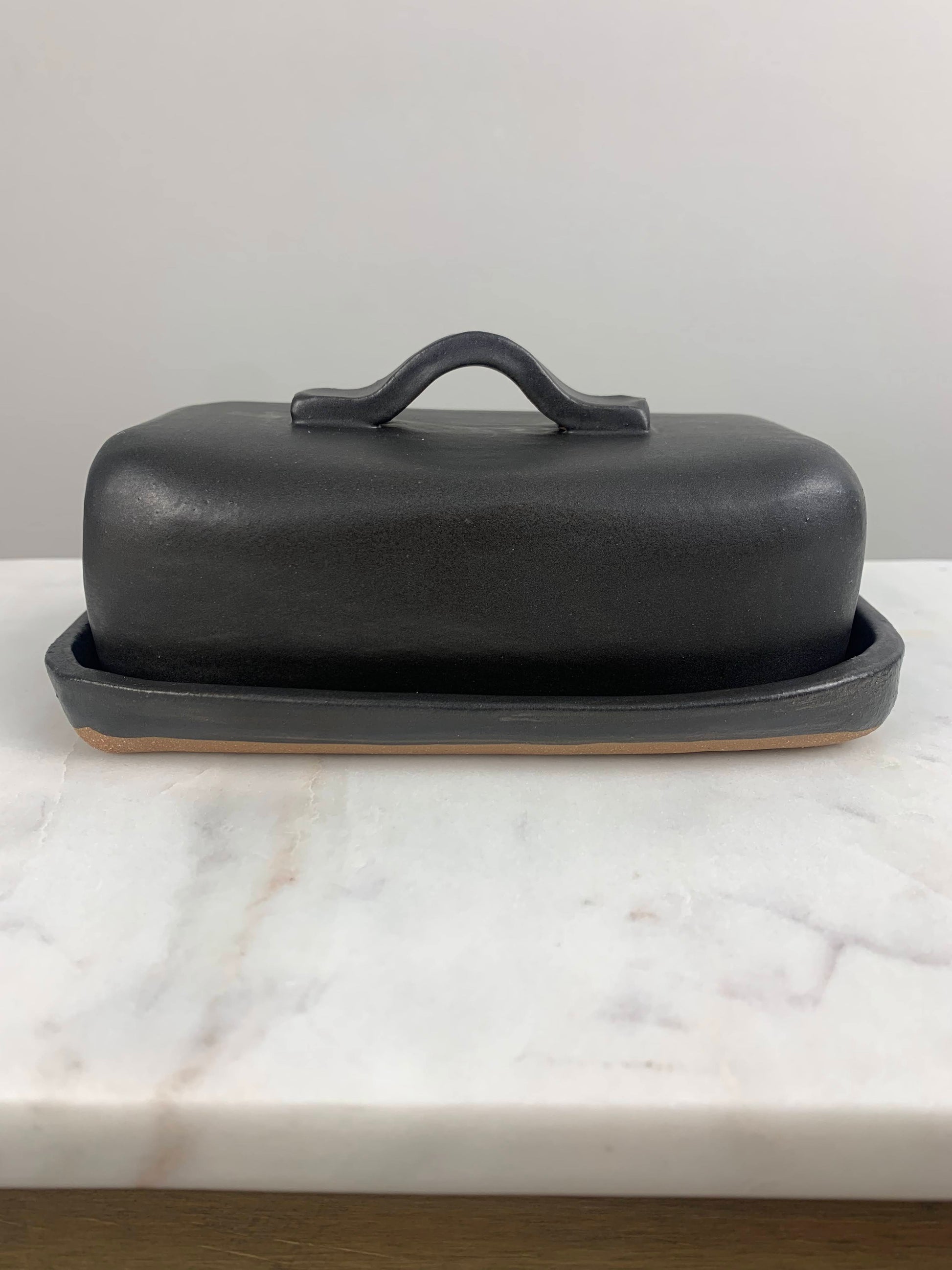 front view of a handcrafted Black ceramic butter dish with lid and simple strap handle on a marble surface