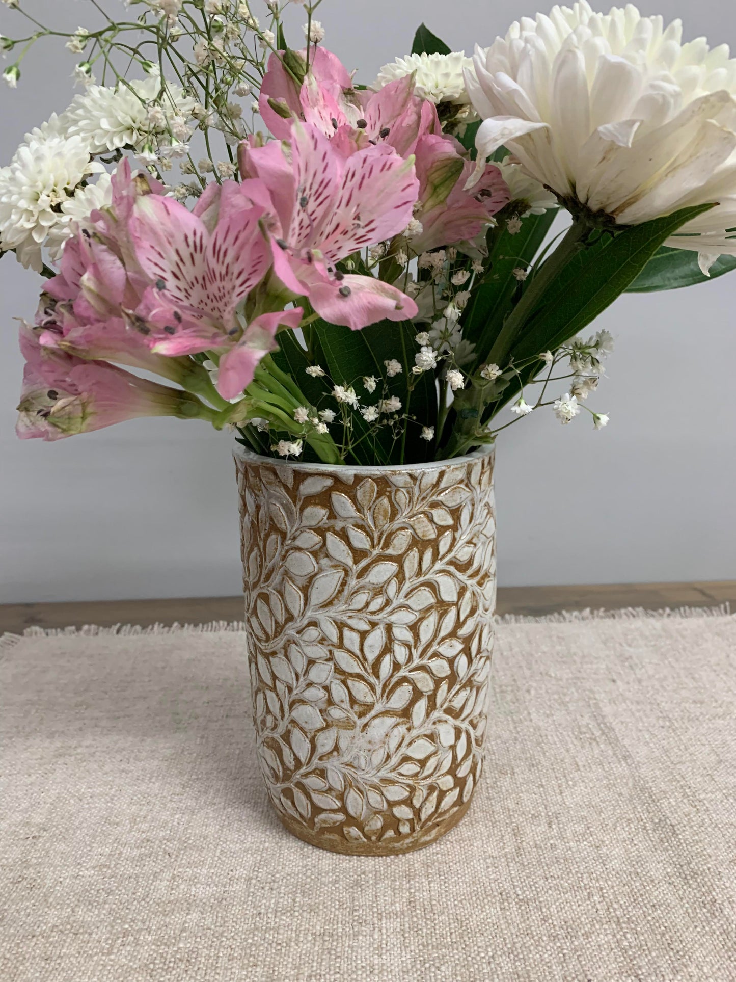 Cylindrical handmade ceramic vase with intricate leaf pattern full of flowers on a textured surface