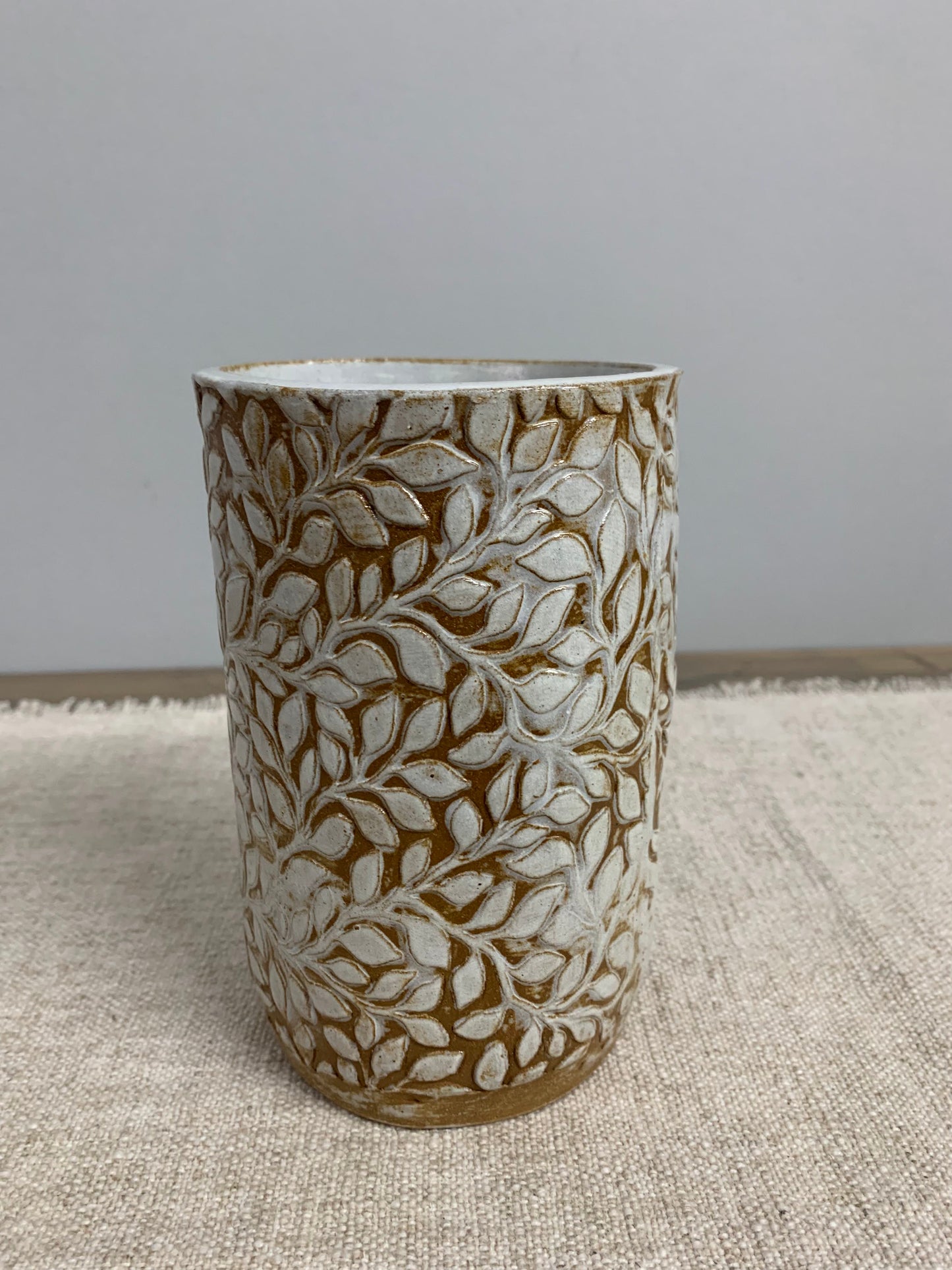 Cylindrical handmade ceramic vase with intricate leaf pattern on a textured surface