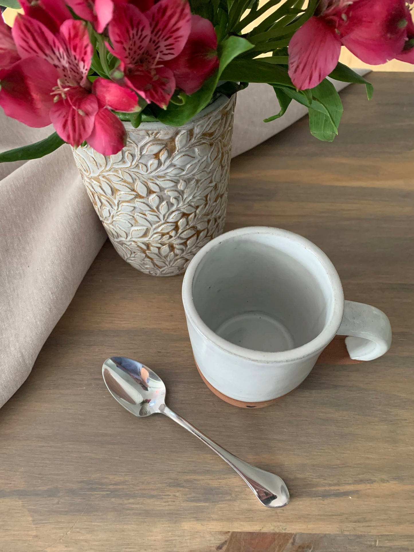 spoon and handcrafted ceramic mug next to a cylindrical vase with embossed leaf designs holding a bouquet of flowers