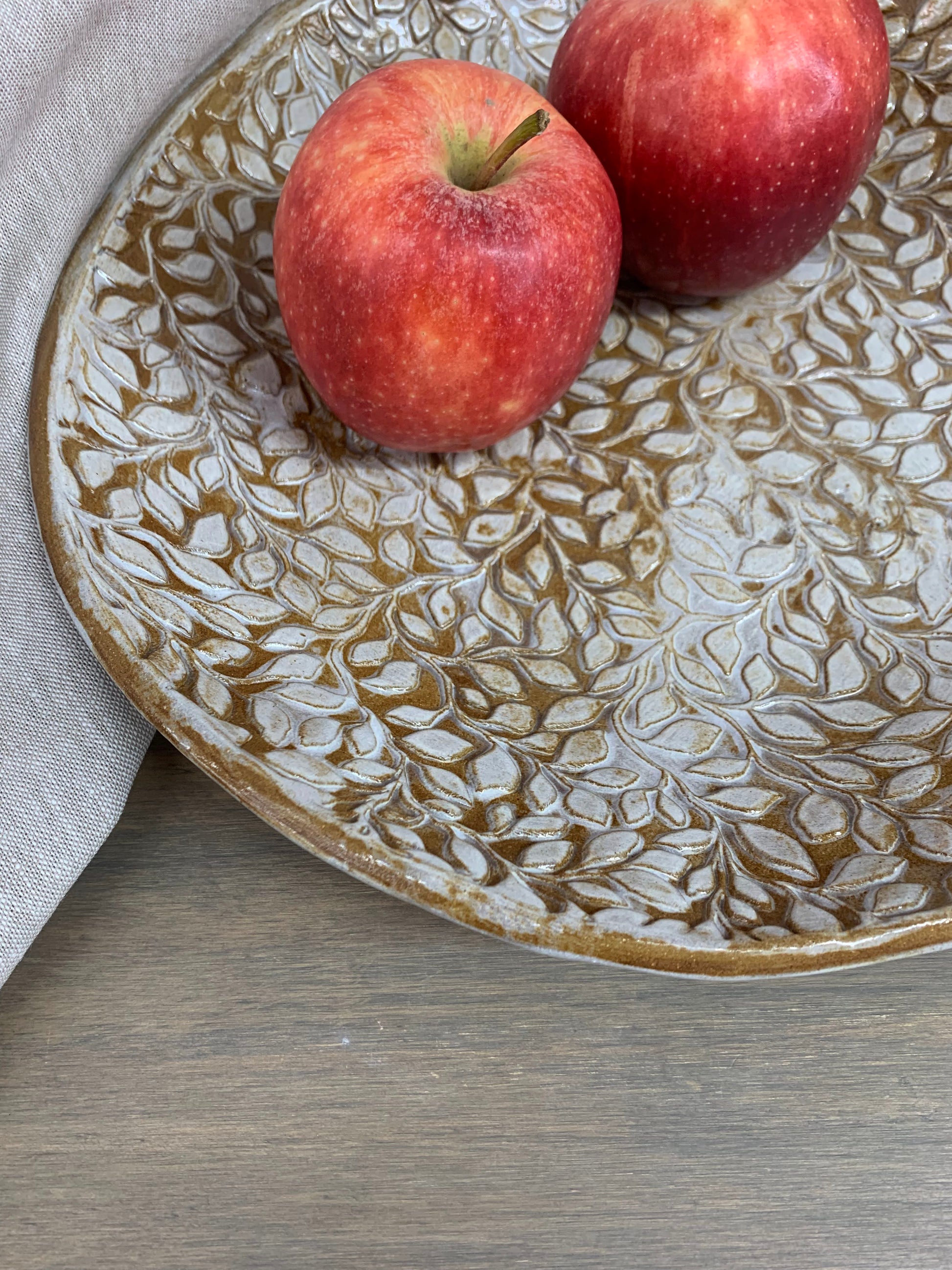 Two red apples on a decorative ceramic plate with a leaf pattern on a wooden surface.